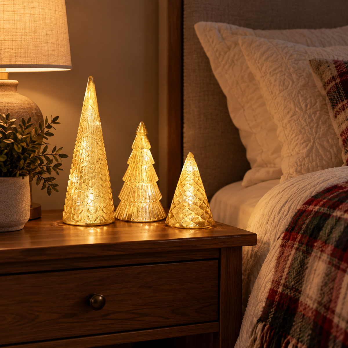 Three gold lighted Christmas trees glowing on a wooden nightstand beside a bed with a plaid throw