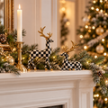 Black and white checkered Christmas deer figurines with gold antlers on a white fireplace mantel with pine garland and a gold candlestick